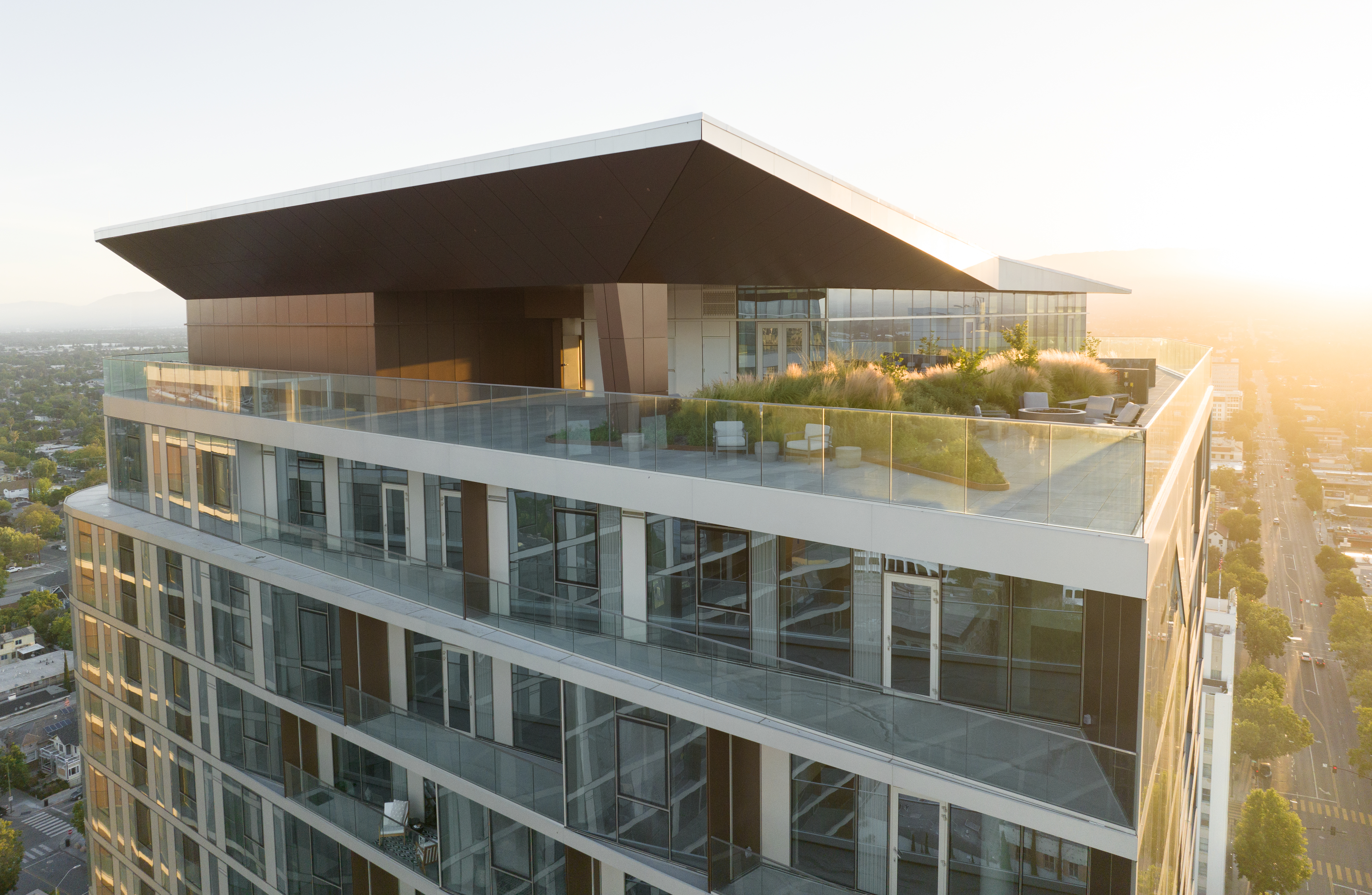 A modern high-rise building with large glass windows and a spacious rooftop terrace featuring plants, seating areas, and city views, bathed in bright sunlight.