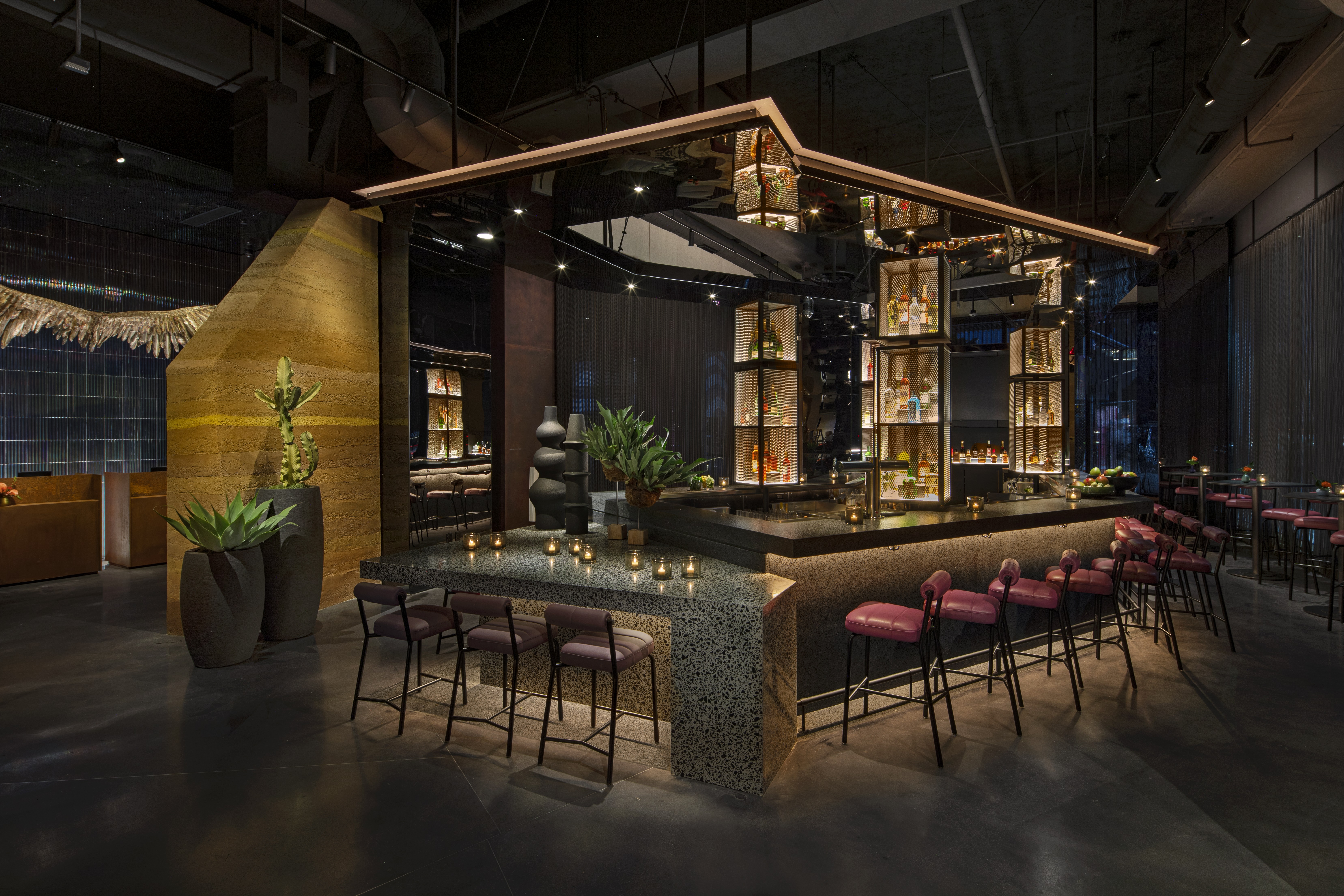 A modern, dimly lit bar with a central granite counter, pink cushioned bar stools, hanging shelves with bottles, potted plants, and candles creating a warm, inviting atmosphere.