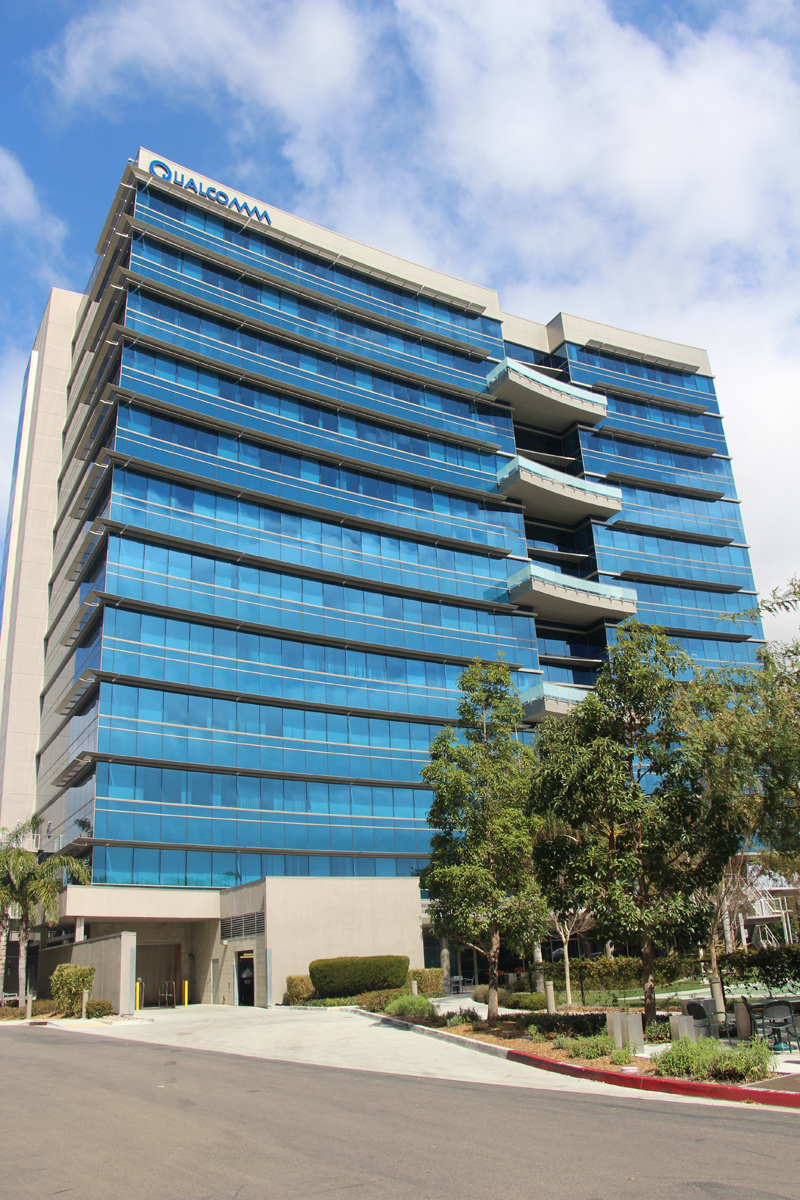 A modern multi-story office building with blue-tinted glass windows and a Qualcomm sign on top, surrounded by trees and a clear, partly cloudy sky.