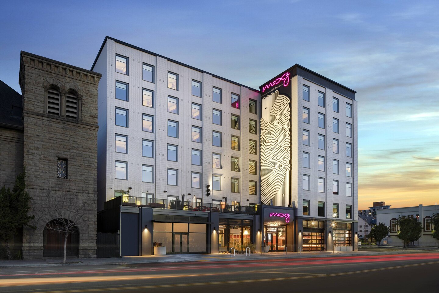 A modern six-story hotel with a lighted Moxy sign and fingerprint design on the facade, next to an older stone building, with cars passing by at dusk and a colorful sky in the background.