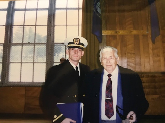 A young man in a naval officer uniform stands next to an older man in a suit and tie, posing indoors in front of large windows with wooden paneling on the wall behind them.