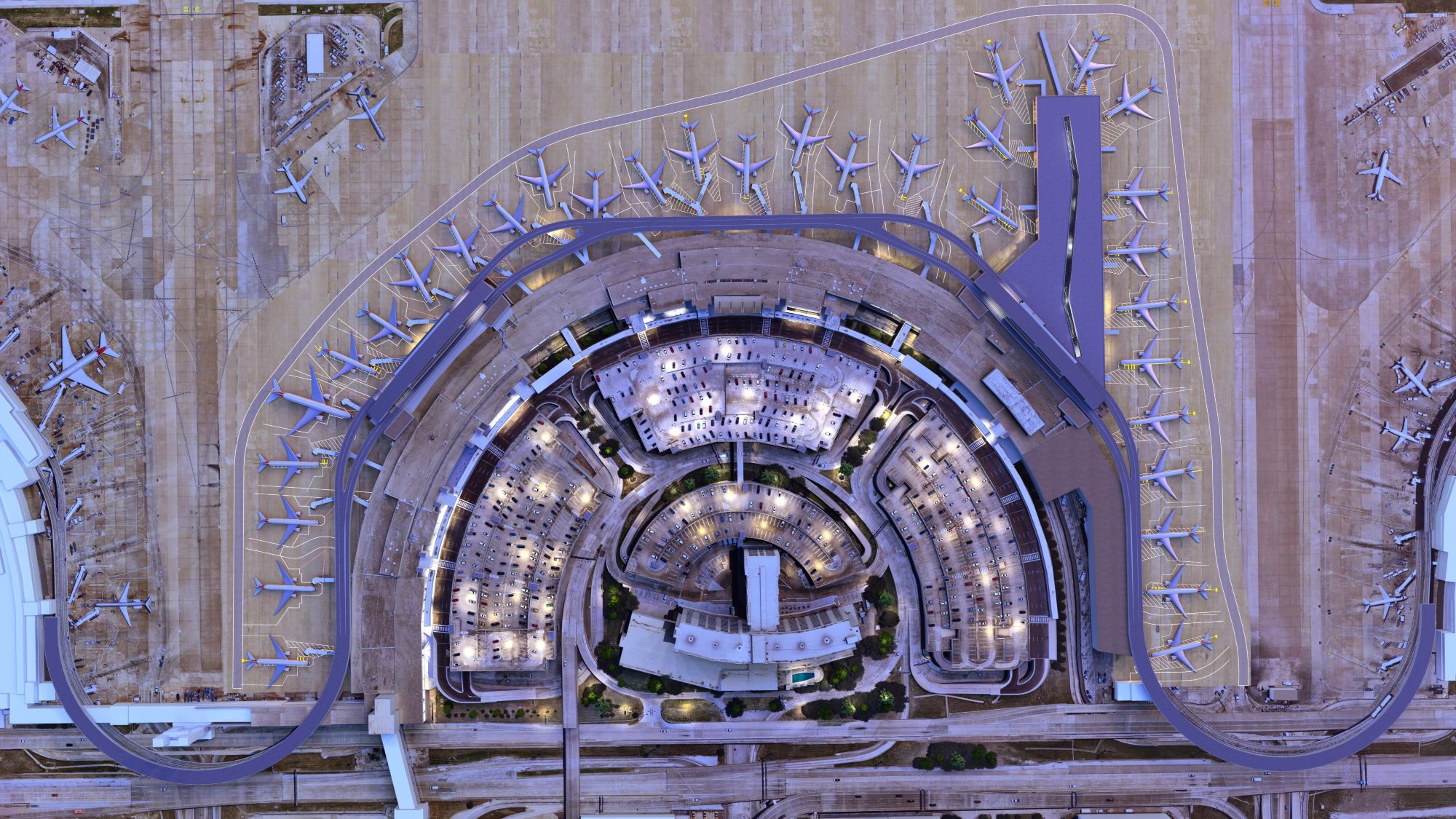 Aerial view of a large airport terminal with multiple airplanes parked at gates around a curved building, surrounded by runways, taxiways, and roads. The terminal is lit with bright lights.