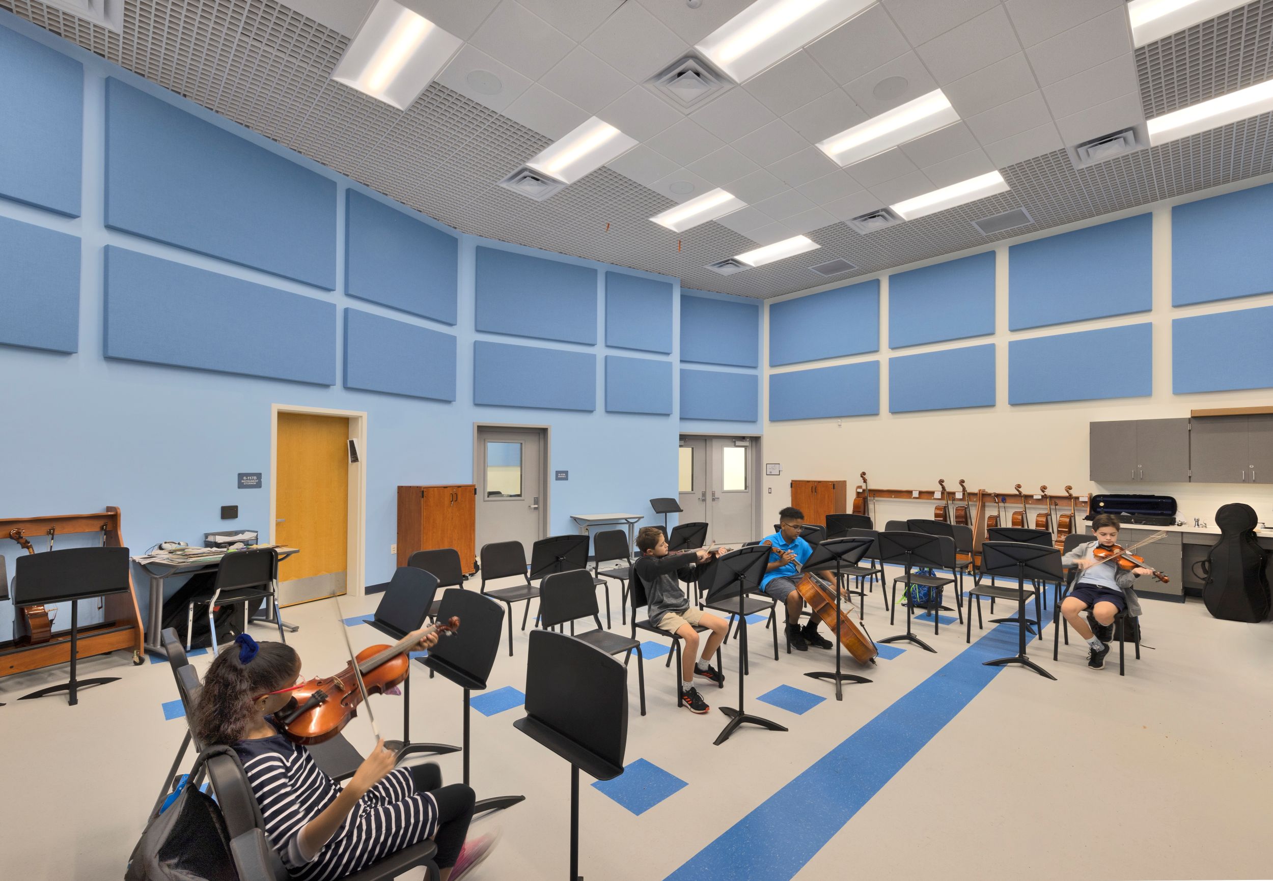 Four children play string instruments in a spacious, modern music classroom with blue soundproof panels, empty music stands, and bright overhead lights.