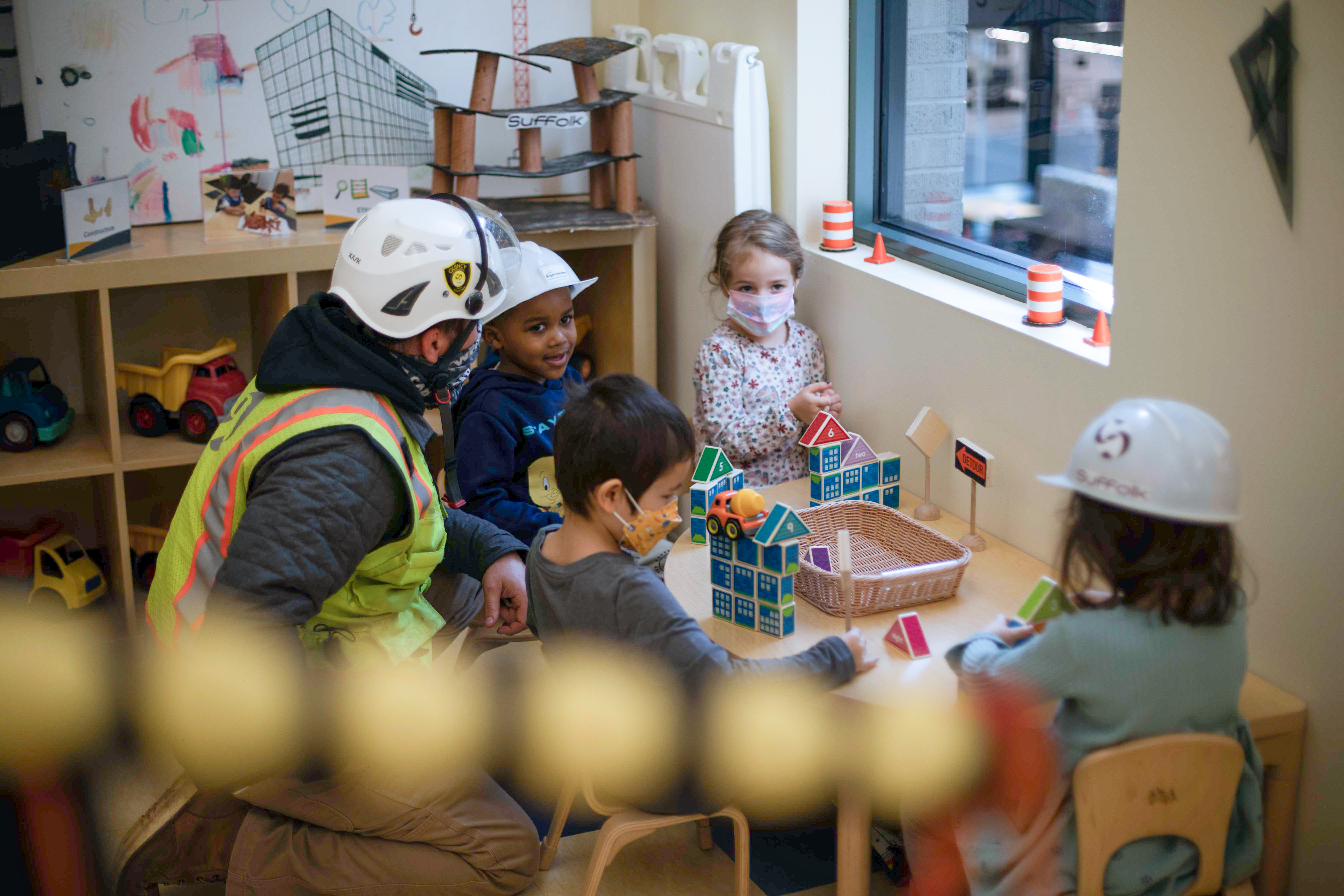 A construction worker and four young children wearing masks and hard hats sit around a table building structures with colorful blocks in a classroom decorated with toy trucks and construction drawings.