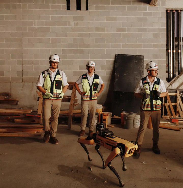 Three construction workers wearing safety vests and helmets stand in a partially built room with scattered materials. In front of them is a yellow, dog-like robot with sensors on its back.