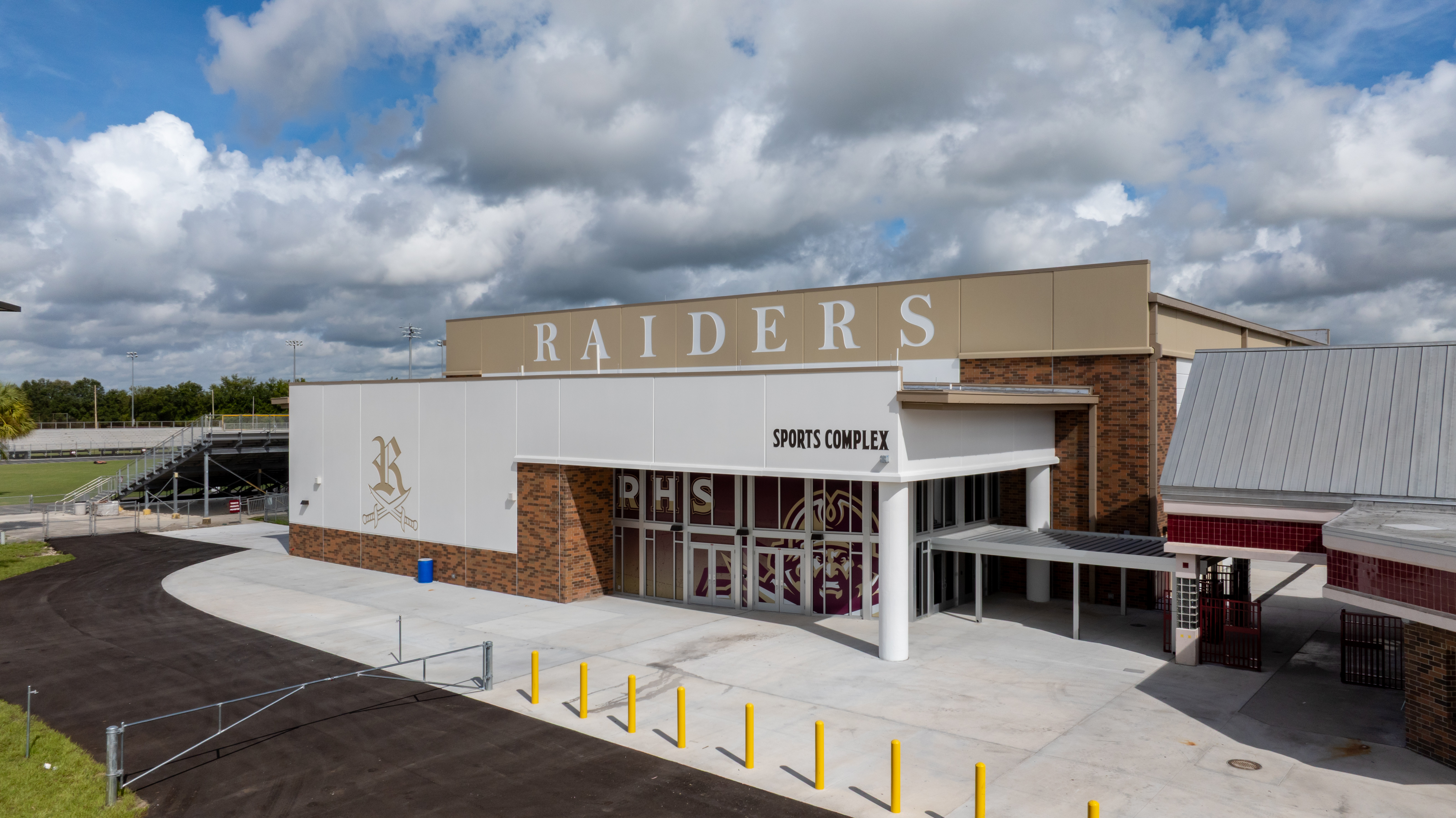 Exterior view of a sports complex building with Raiders written on top and school initials on the glass doors, under a partly cloudy sky. Yellow posts and a paved walkway are in front of the entrance.