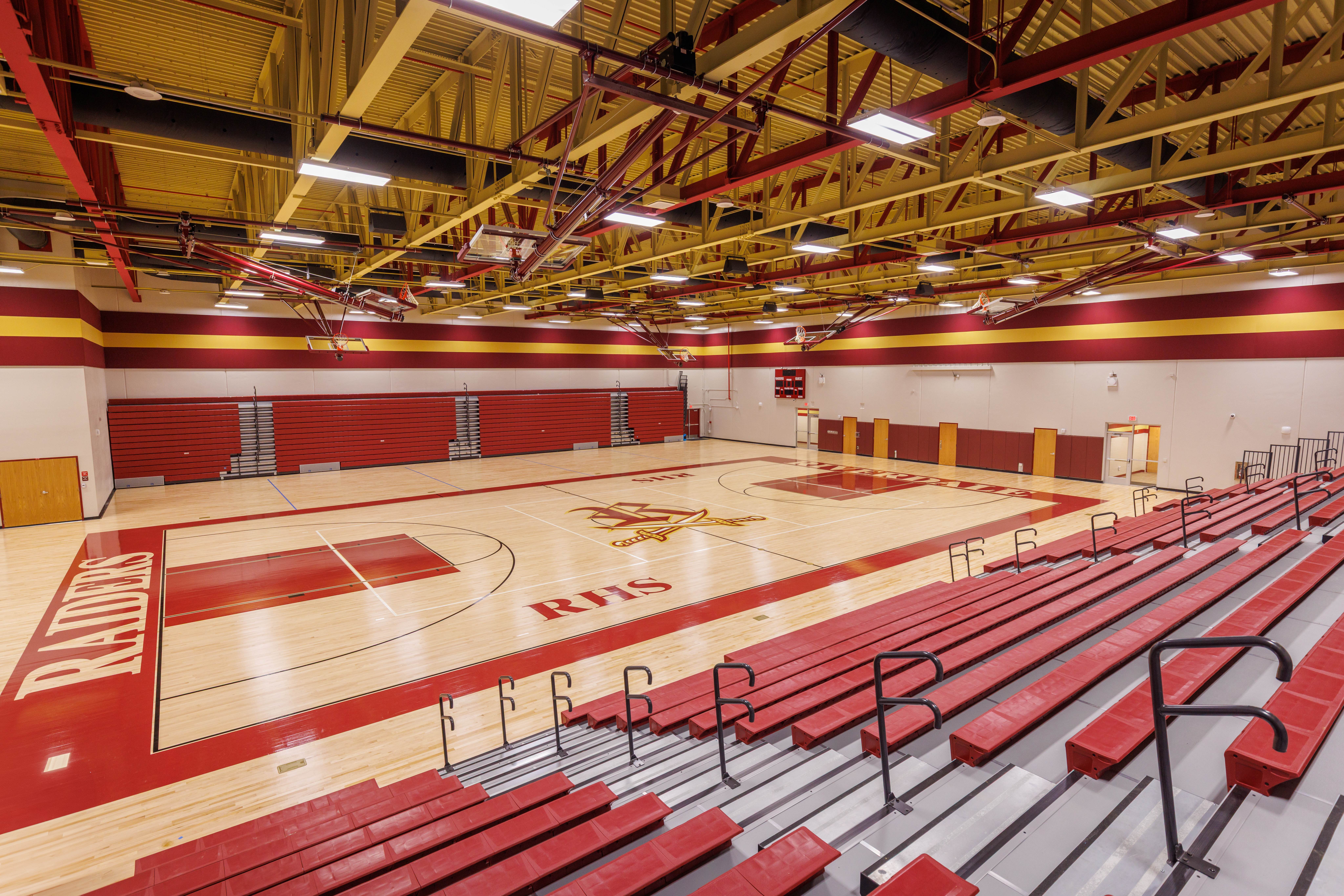 Wide view of a spacious indoor gymnasium with red and yellow accents, wooden floors featuring a basketball court, bleachers on one side, and the word “RAIDERS” painted along one baseline.