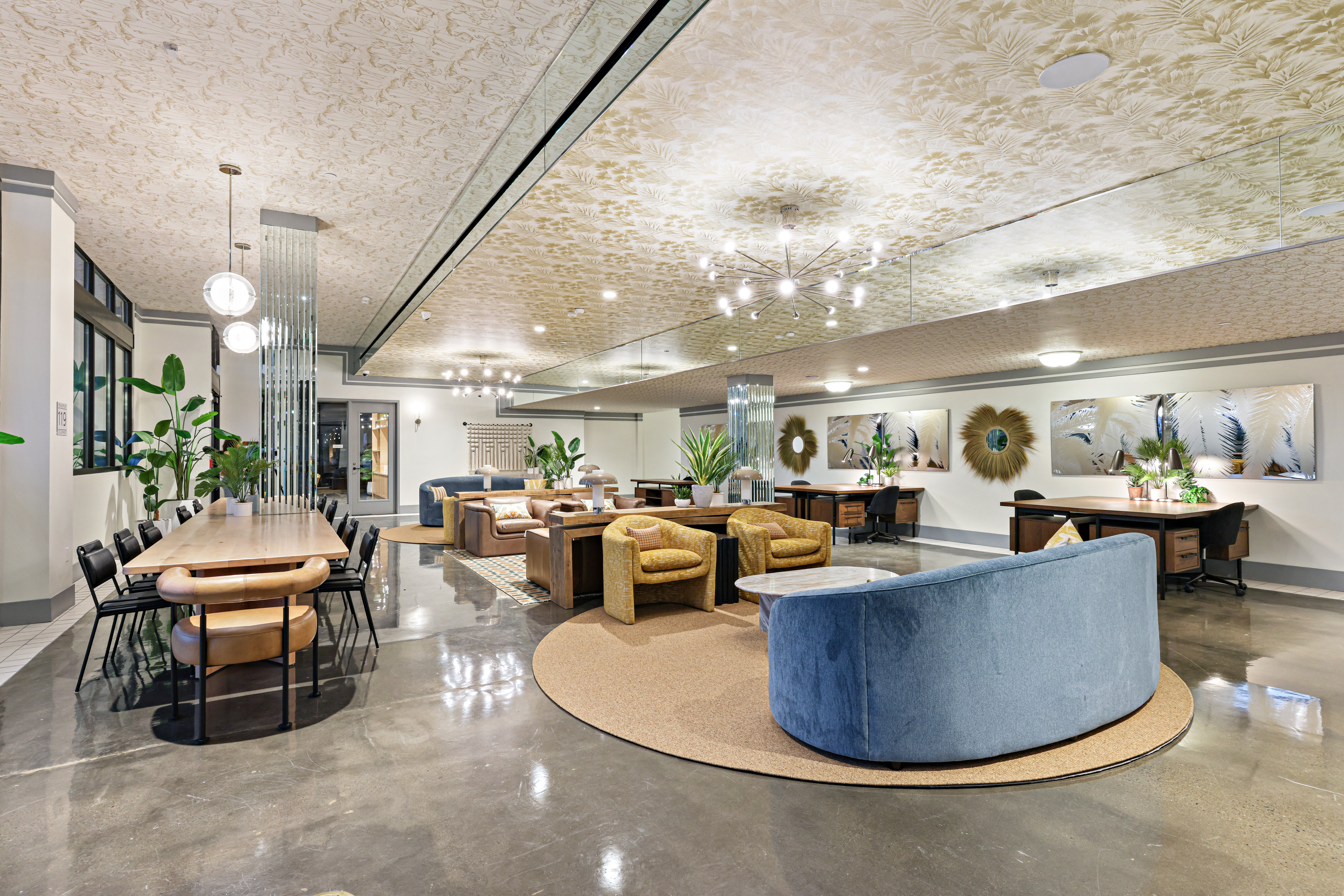 Modern, spacious lounge with glossy floors, a long wooden table with chairs, cozy armchairs, sofas, indoor plants, decorative mirrors, and framed artwork. Soft lighting and patterned ceiling create a welcoming atmosphere.