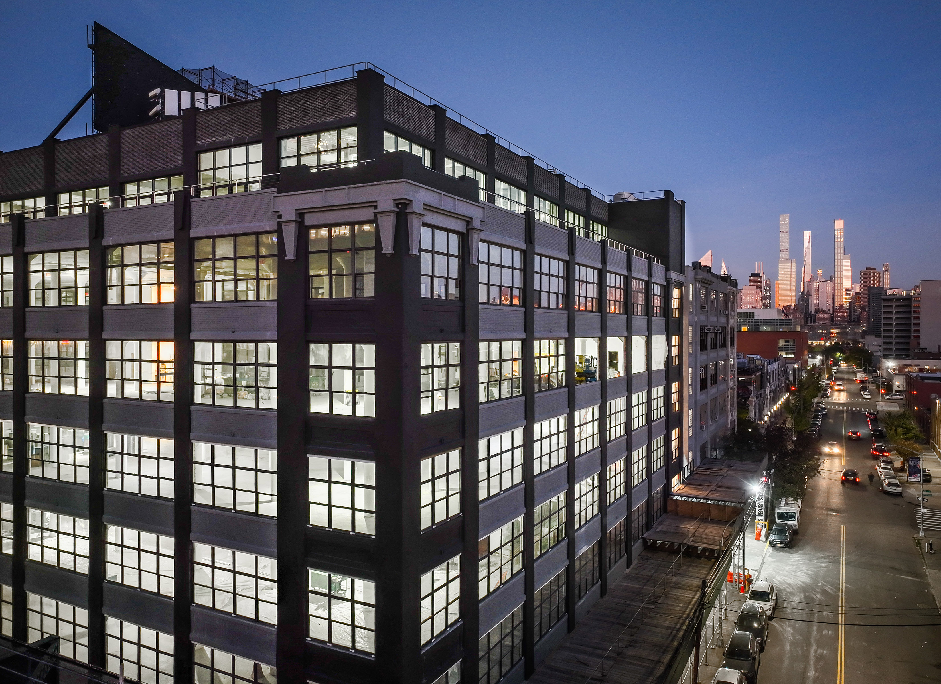 A large, multi-story office building with many illuminated windows is shown at dusk, with a city street and tall skyscrapers visible in the background under a darkening sky.