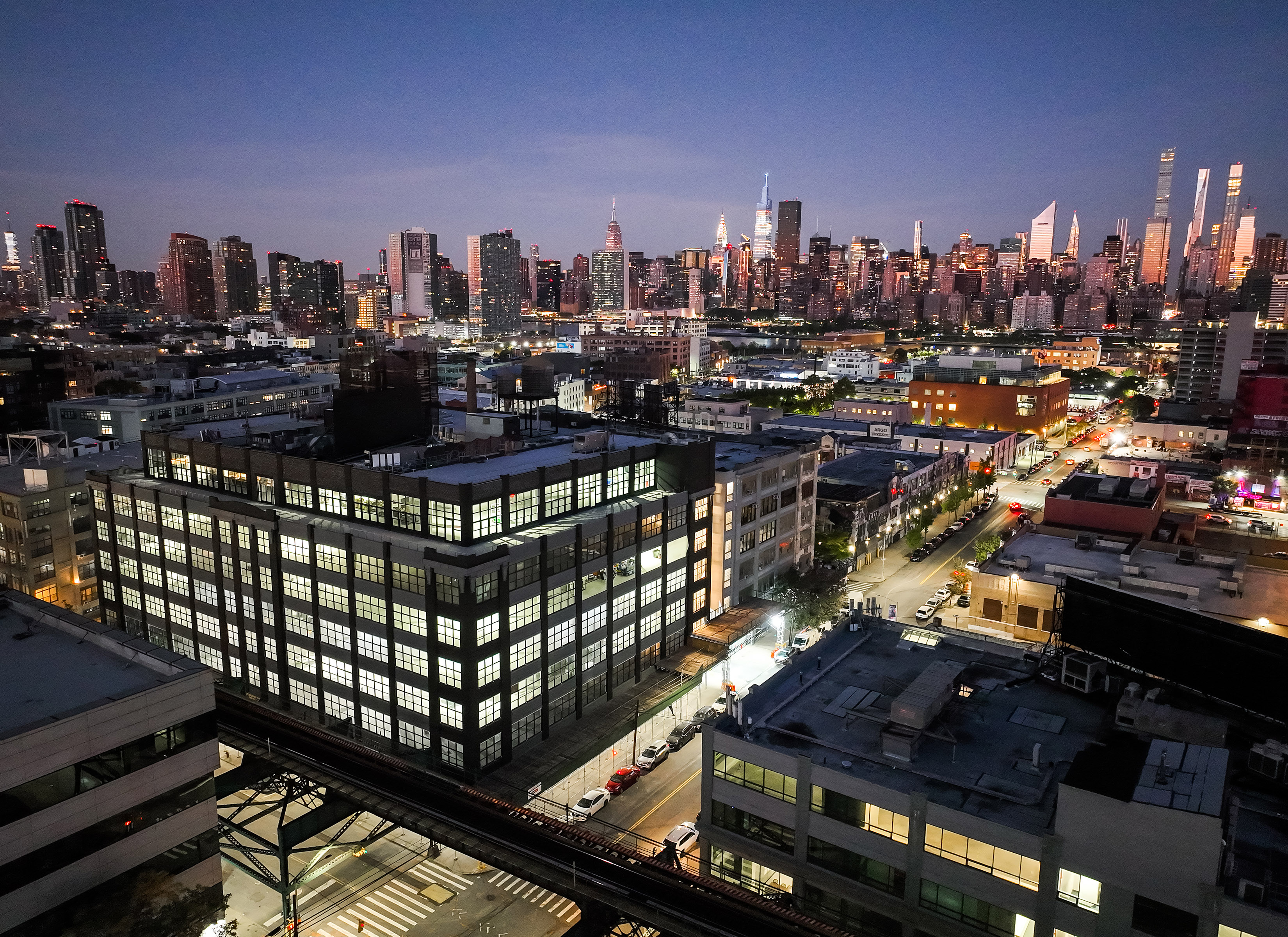 A cityscape at dusk shows an illuminated office building in the foreground, busy streets below, and a skyline of tall skyscrapers with lights in the background under a darkening sky.