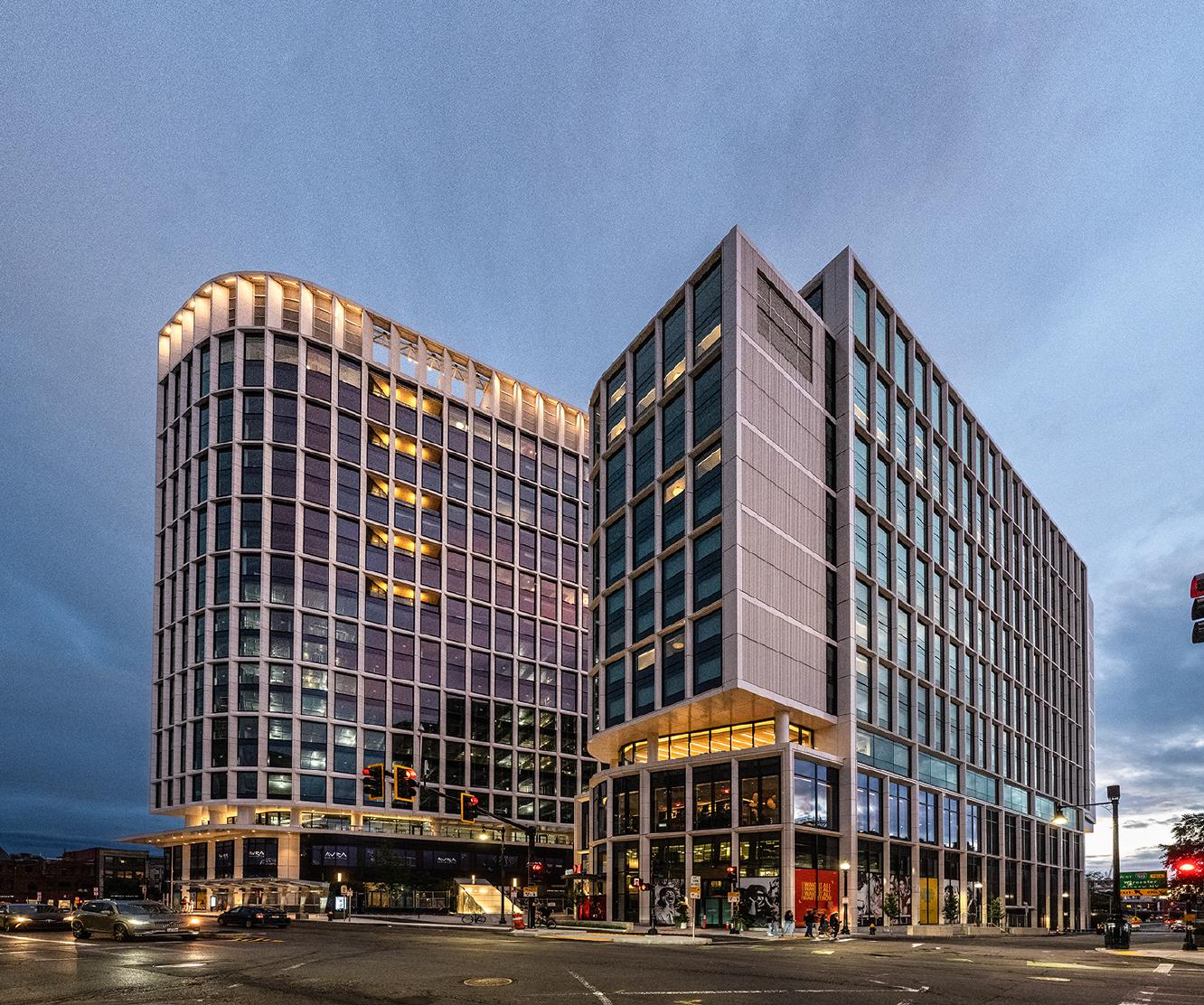 Modern office buildings with large glass windows and illuminated interiors stand at a street corner during dusk, with pedestrians and cyclists passing by on the sidewalk.