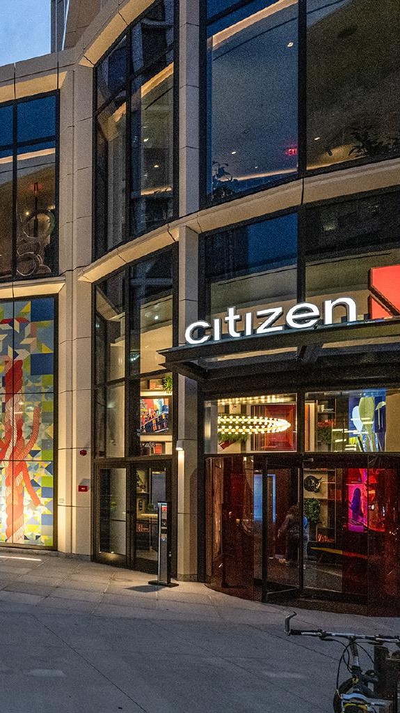 Street view of the entrance to citizenM hotel at dusk, with glass windows, illuminated sign, modern decor inside, a colorful mural on the wall, and a bicycle parked nearby.