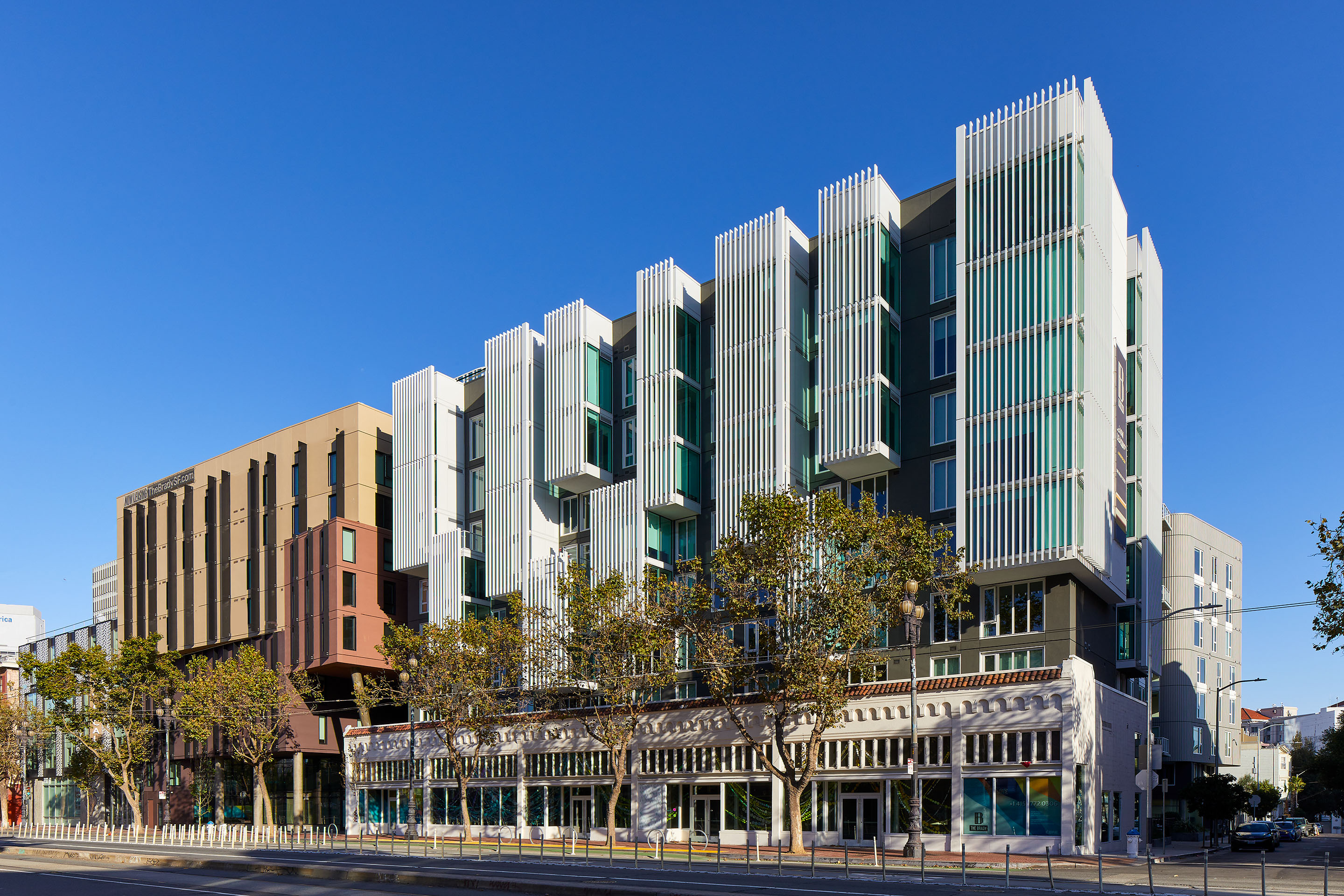 A modern multi-story building with glass façades and vertical white fins, standing above older-style storefronts, with trees lining the street and a clear blue sky in the background.