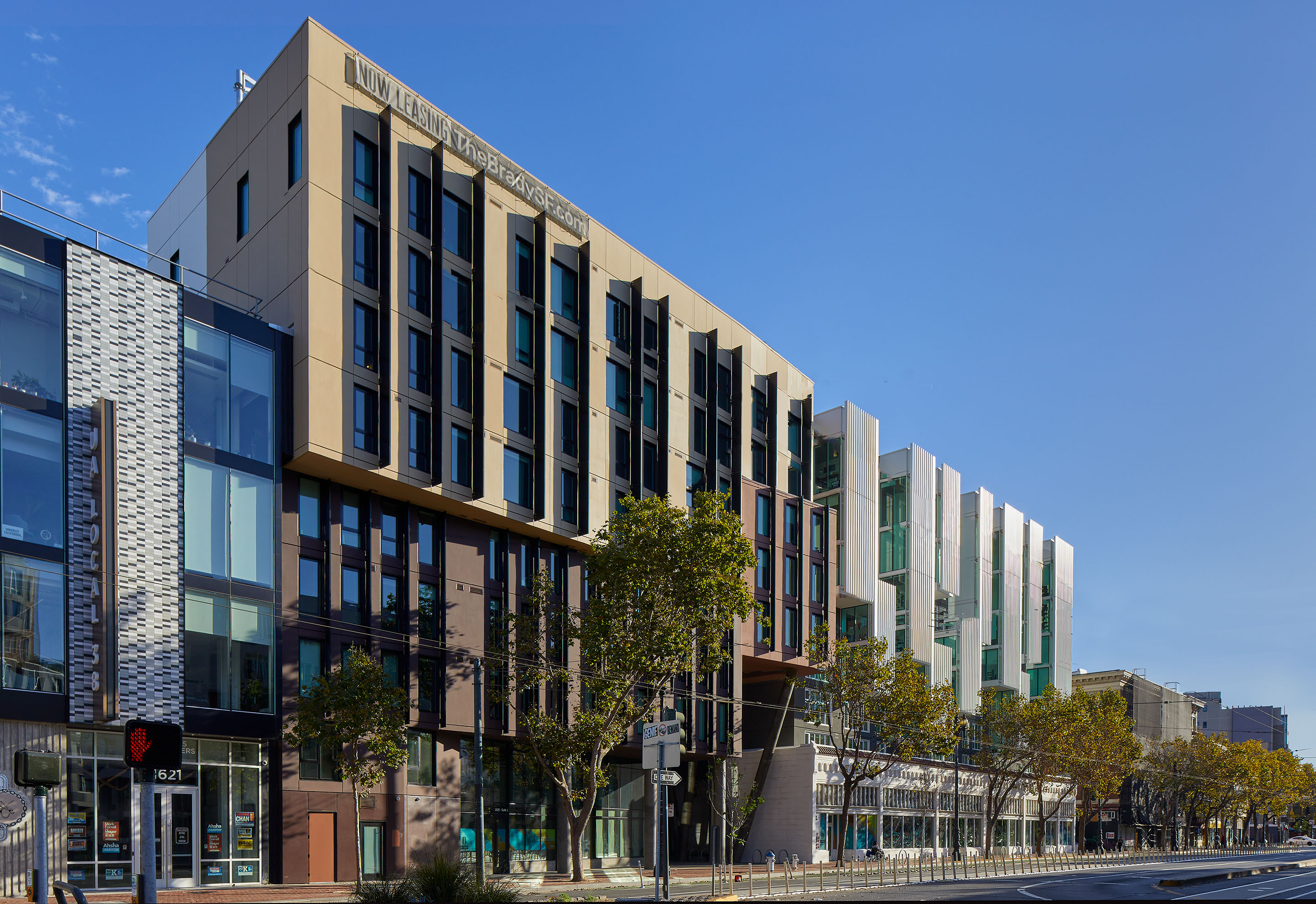 A modern multi-story building with large windows and varied facade materials stands beside a tree-lined street on a clear, sunny day. Nearby, other contemporary structures and shops are visible.
