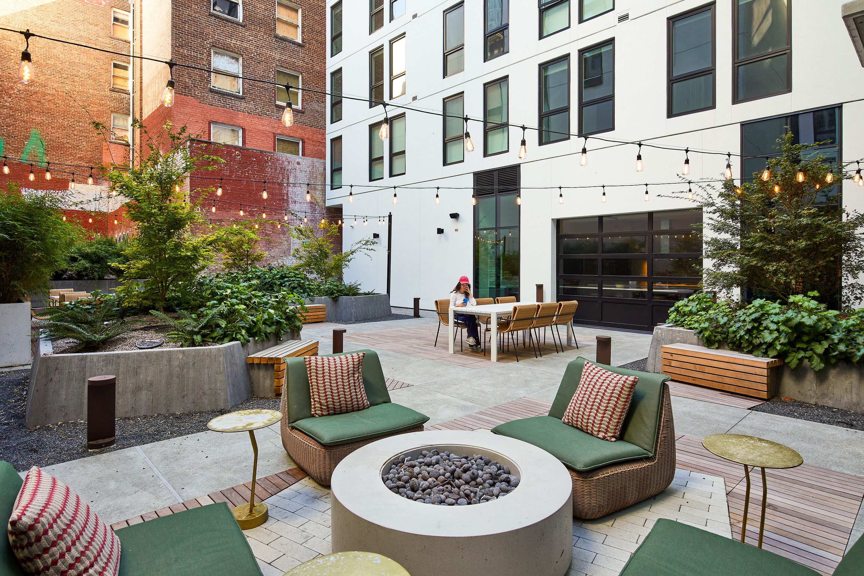 A modern outdoor patio with green cushioned chairs around a fire pit, string lights overhead, and a long dining table; one person sits at the table, surrounded by plants and city buildings.