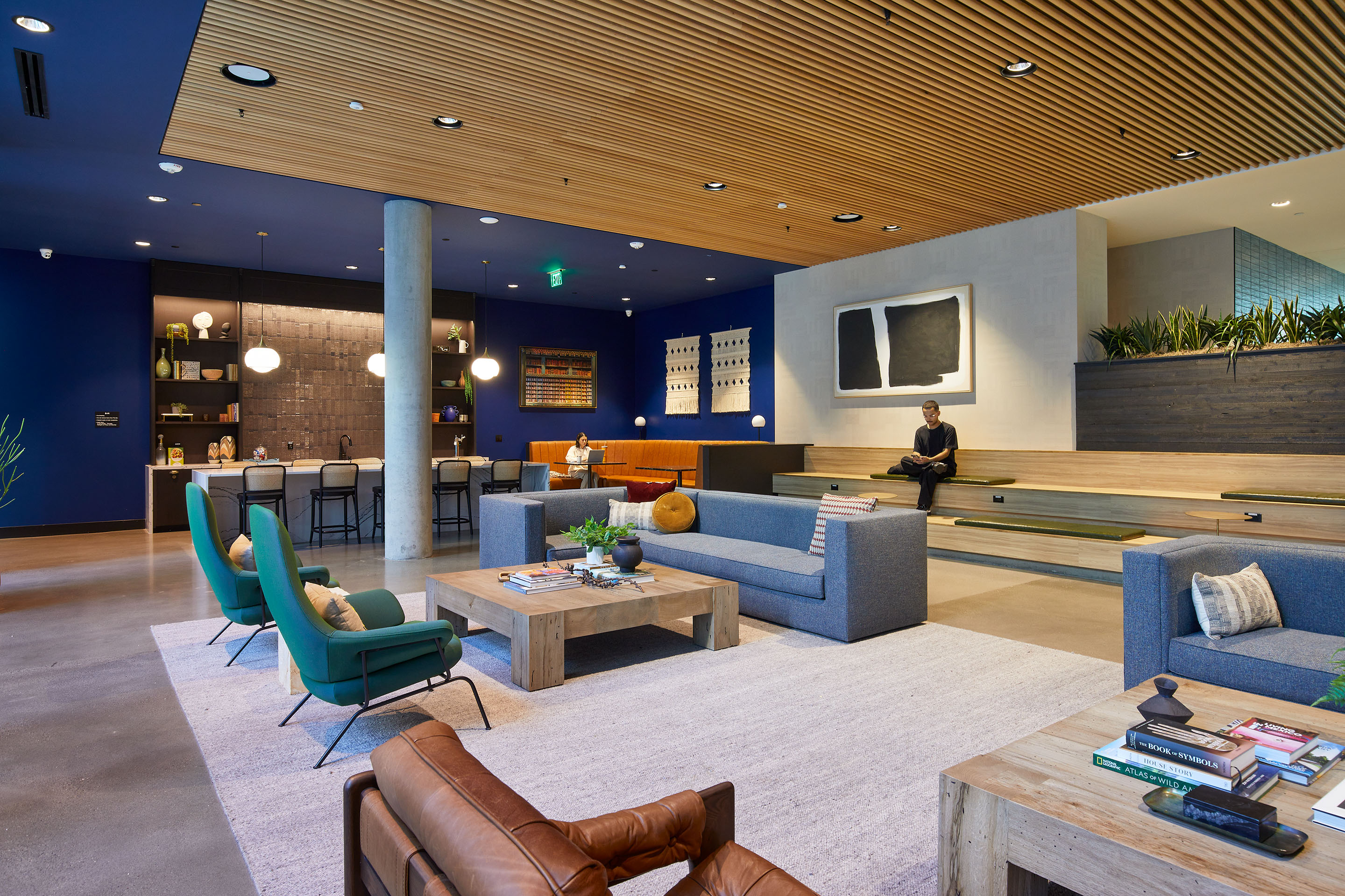 A modern lounge with blue walls, comfy seating, a wooden ceiling, a bar area with stools, books and decor on tables, plants, and a person sitting on bench seating along one wall.