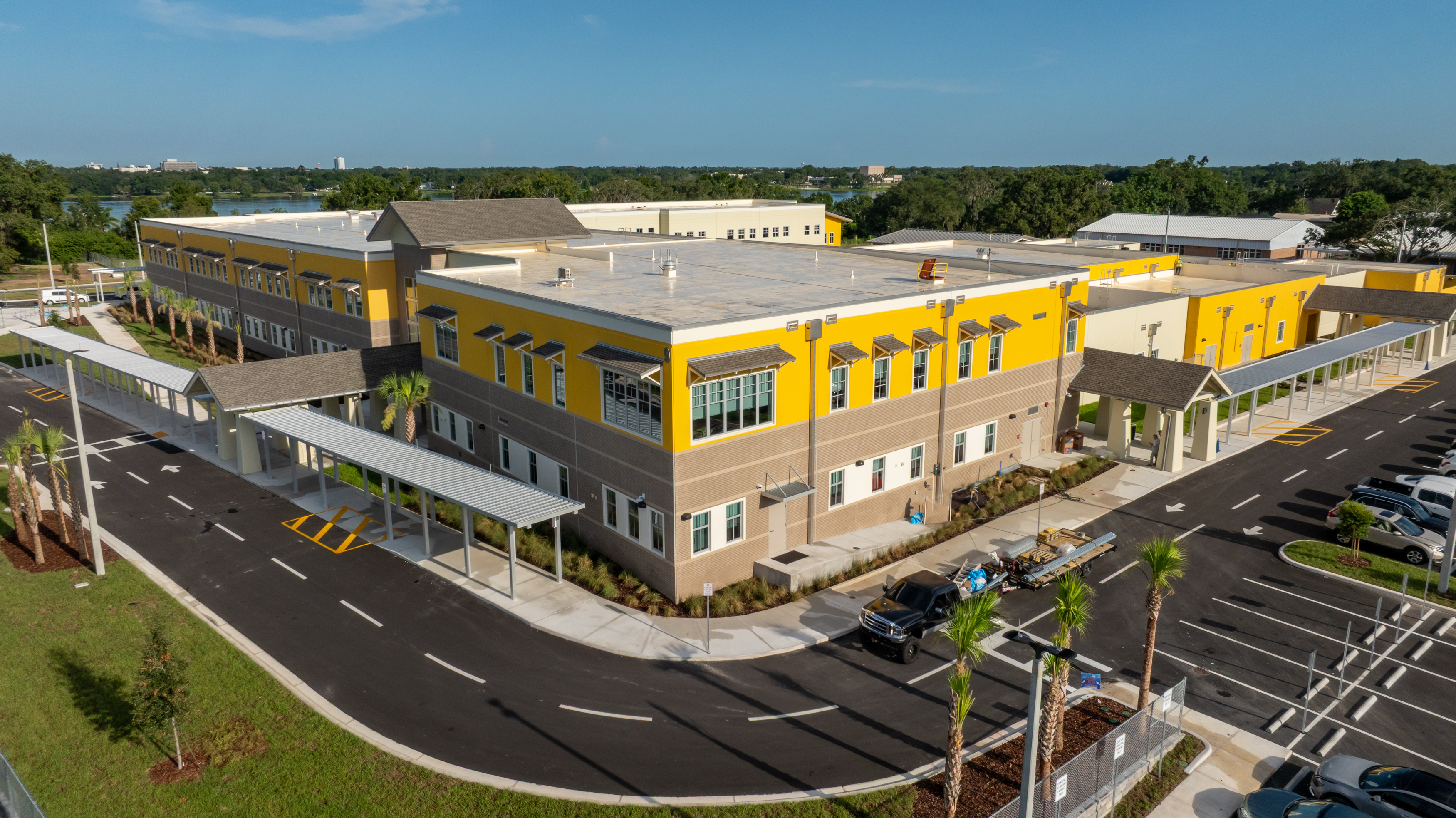 A modern, two-story school building with yellow and beige walls, large windows, covered walkways, and a parking lot surrounded by palm trees under a clear blue sky.