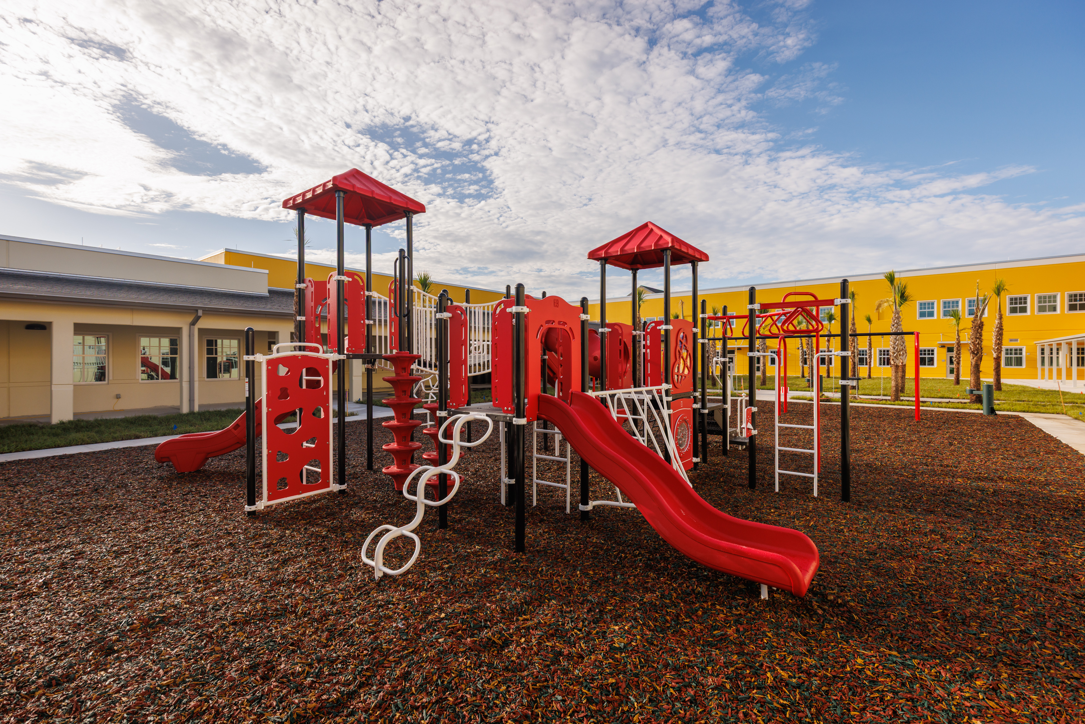 A modern playground with red slides, climbing structures, and ladders sits on a rubber mulch surface, surrounded by yellow buildings under a blue sky with scattered clouds.