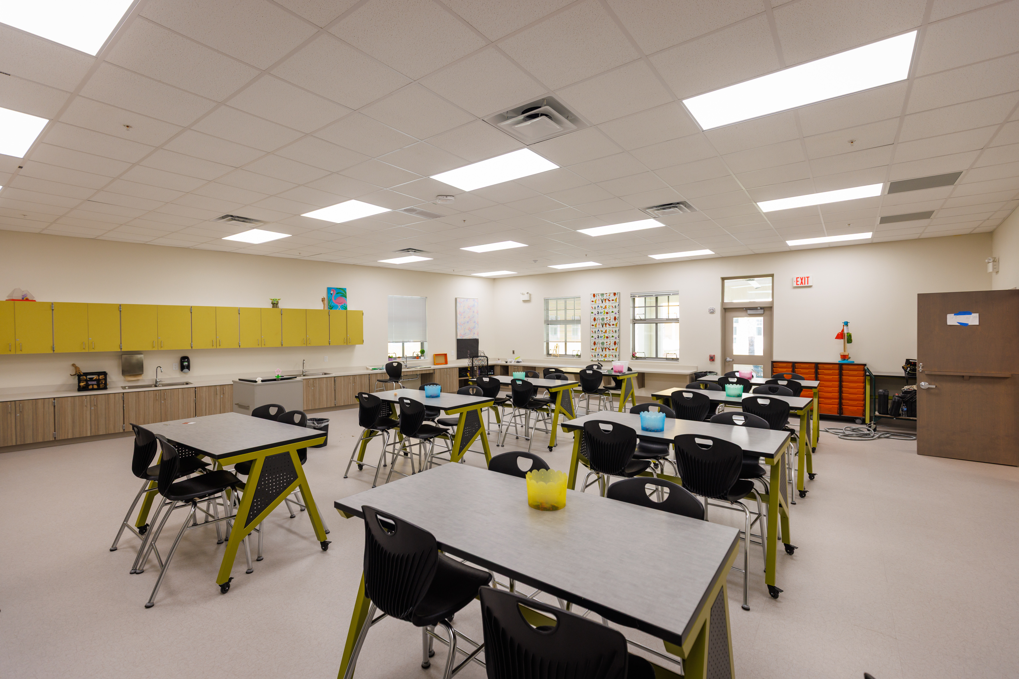 A bright, modern classroom with several gray tables and black chairs. Yellow storage cabinets line the wall, and educational materials and bins are placed around the room. Large windows let in natural light.