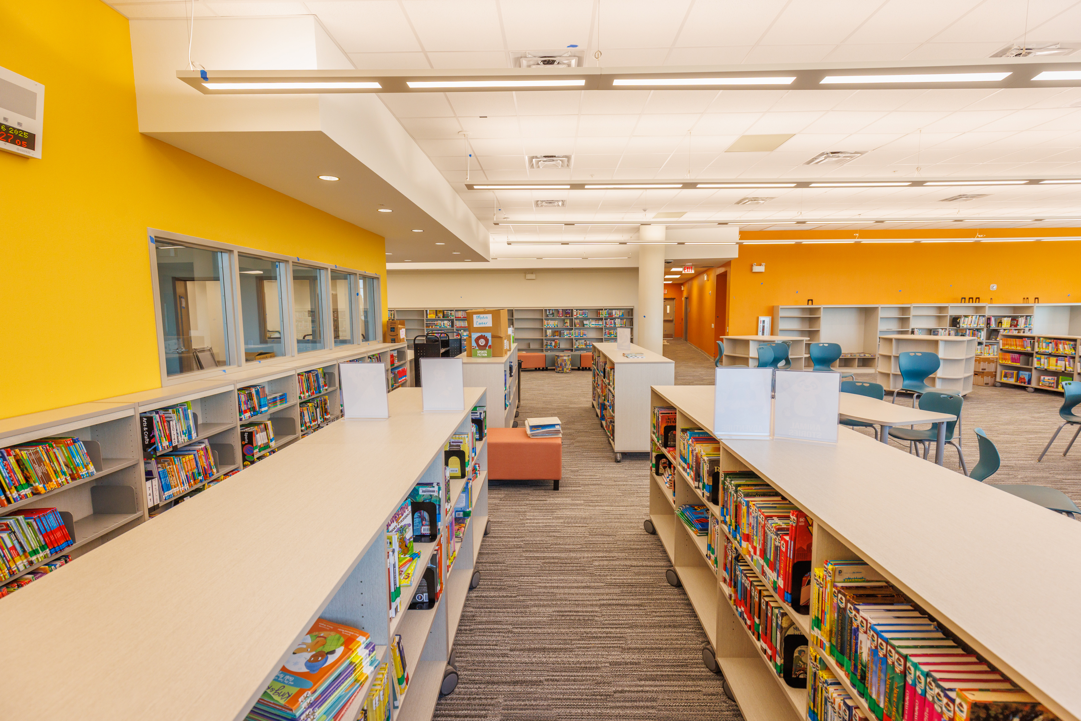 Bright, modern library with yellow walls, carpeted floors, bookshelves filled with colorful books, large windows, and blue chairs arranged around round tables. The space appears spacious and well-lit.