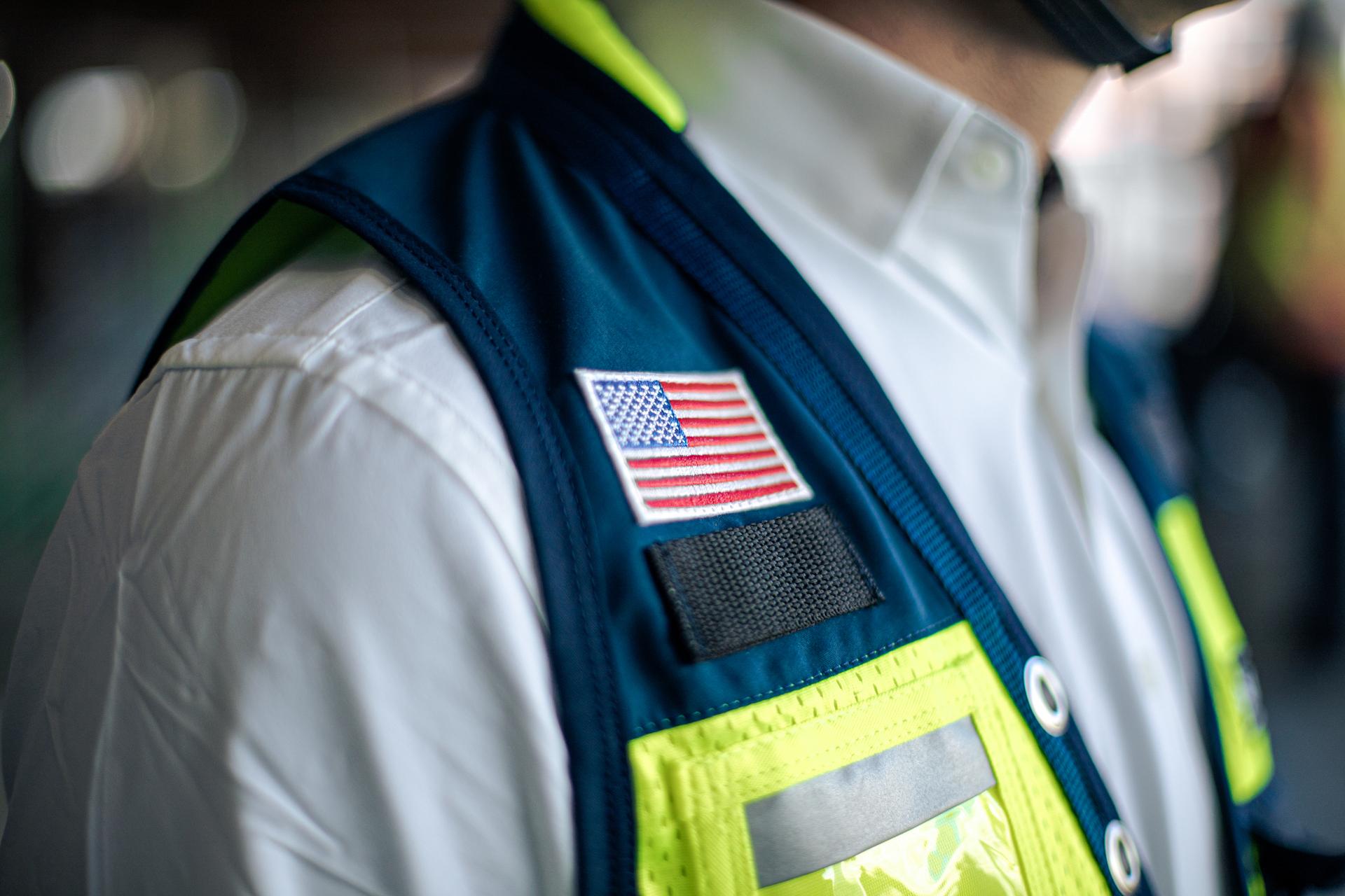 Close-up of a person wearing a white shirt and a dark safety vest with neon yellow stripes, an American flag patch on the shoulder—ready to tackle challenging projects. The person’s face is not visible.