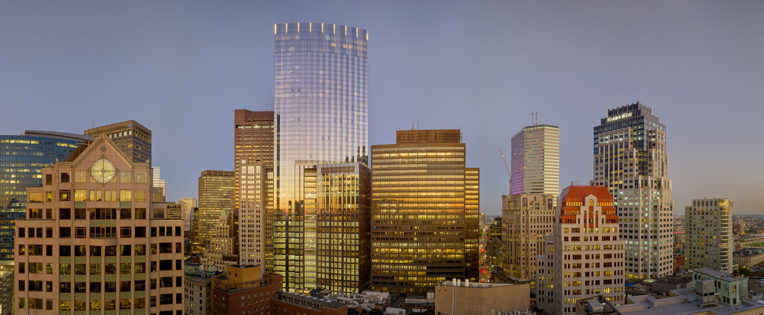 Panoramic view of a city skyline at sunset, featuring modern skyscrapers and office buildings with glass and brick exteriors reflecting the warm golden light.