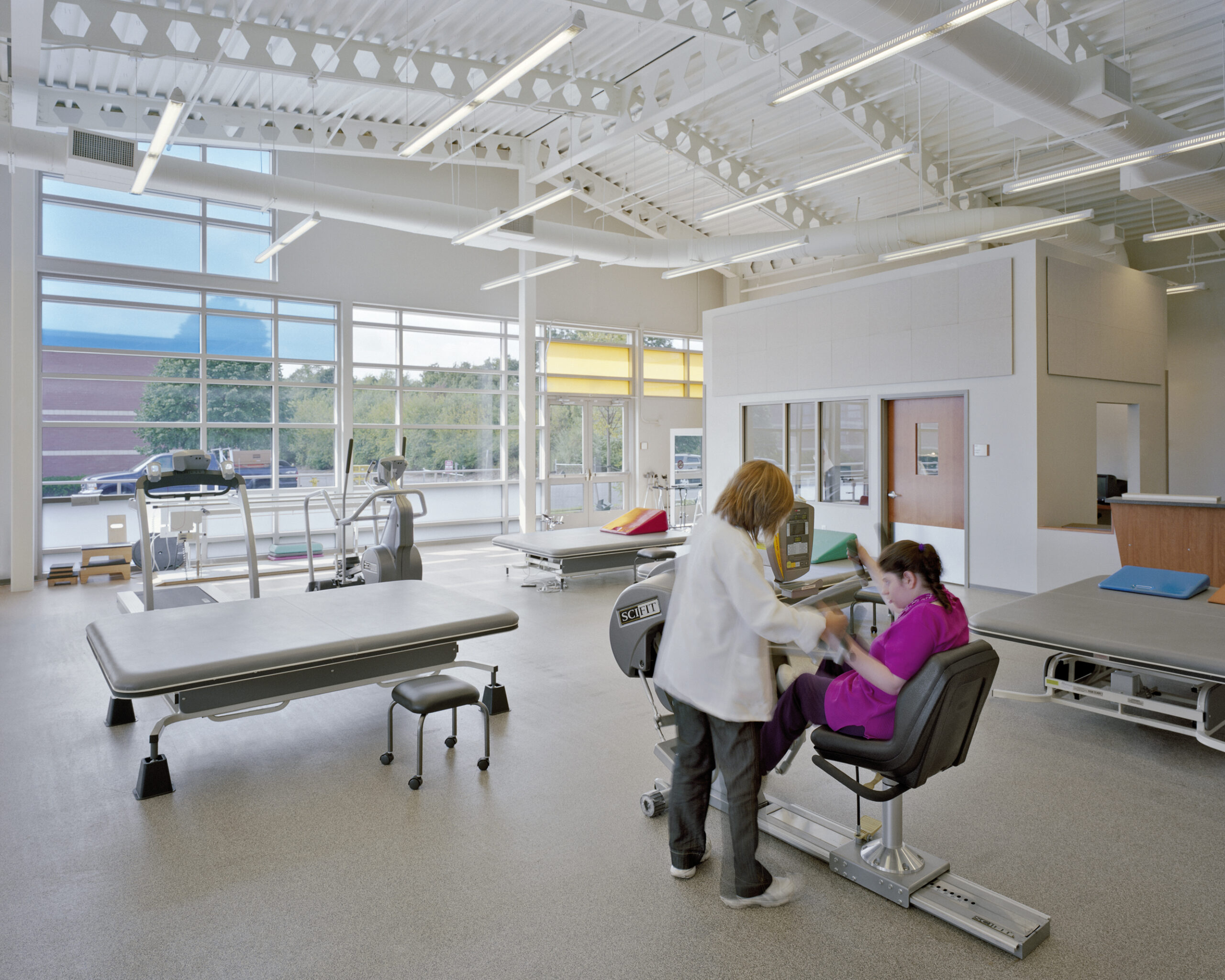 A physical therapist assists a woman seated on exercise equipment in a bright, spacious assisted living rehabilitation center with large windows, therapy tables, and exercise machines.
