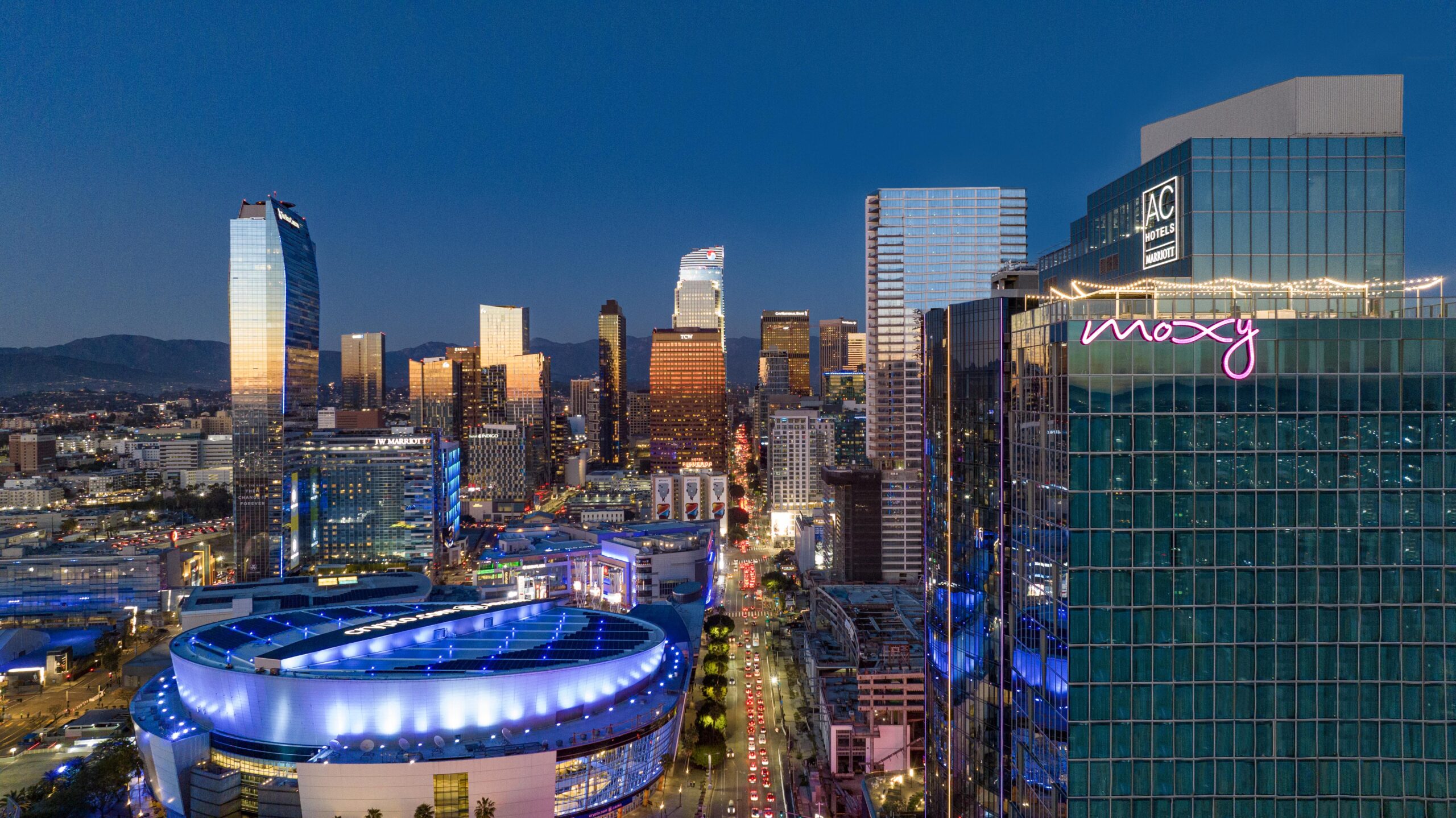 Aerial view of downtown Los Angeles at dusk, featuring illuminated skyscrapers, the Moxy hotel with neon sign, and the round, brightly-lit Crypto.com Arena. City lights and traffic line the main street below.