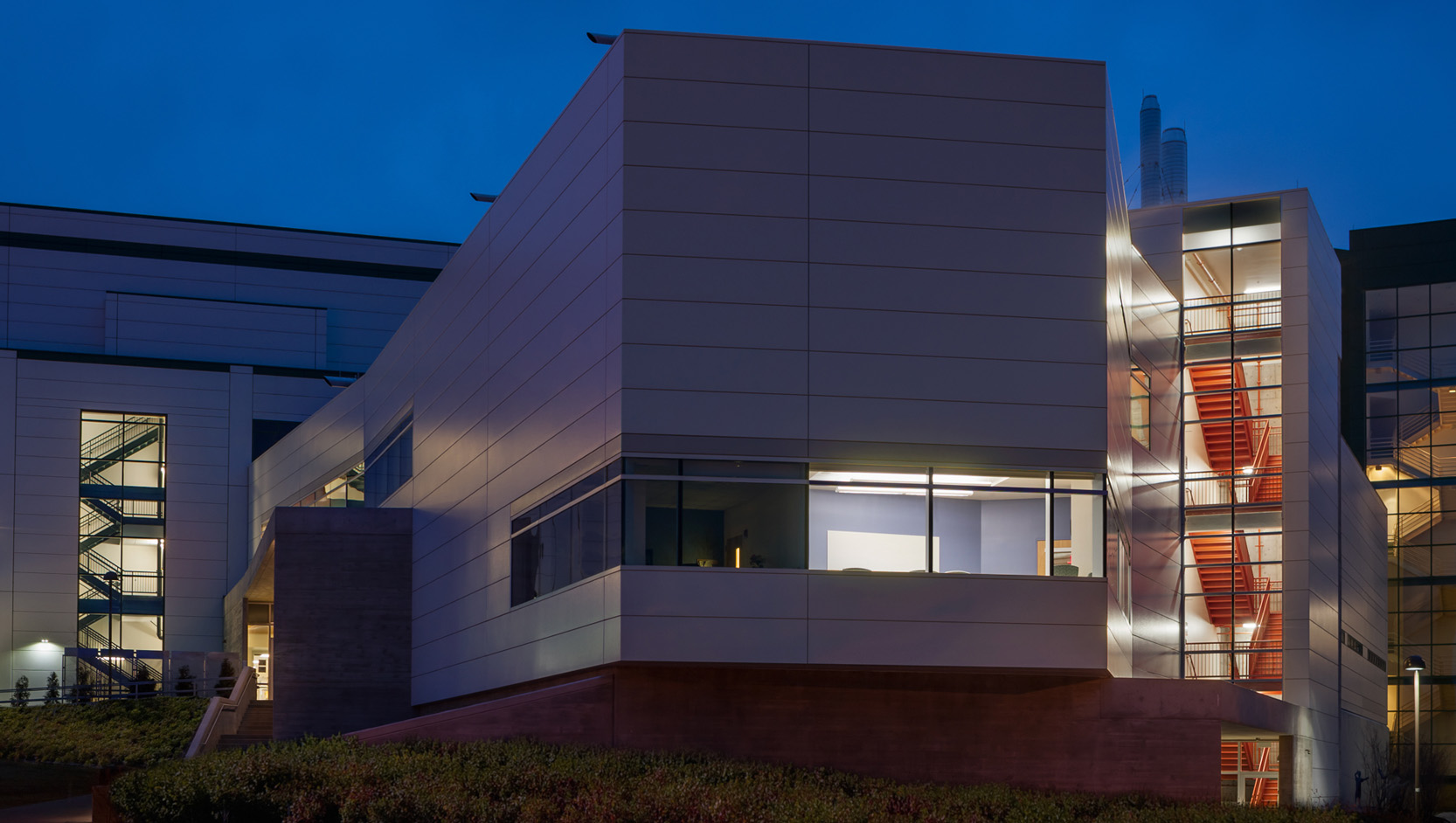 Modern building at dusk with illuminated interior, large windows, and an exterior staircase with red steps, surrounded by greenery and set against a deep blue sky.