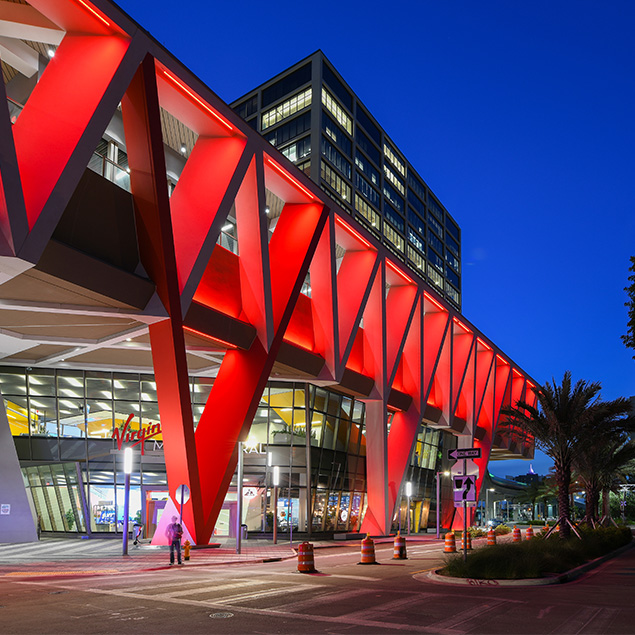 A modern building with bold red geometric supports is brightly lit at night. Large windows reveal the interior, and a high-rise structure is visible above. Palm trees and street signs line the sidewalk.
