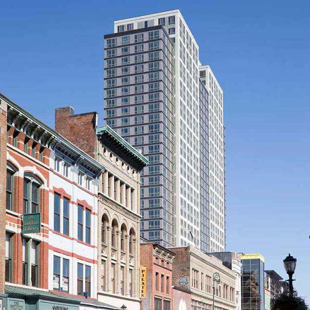 A tall modern high-rise building stands behind a row of historic, low-rise brick buildings with arched windows and colorful facades under a clear blue sky.