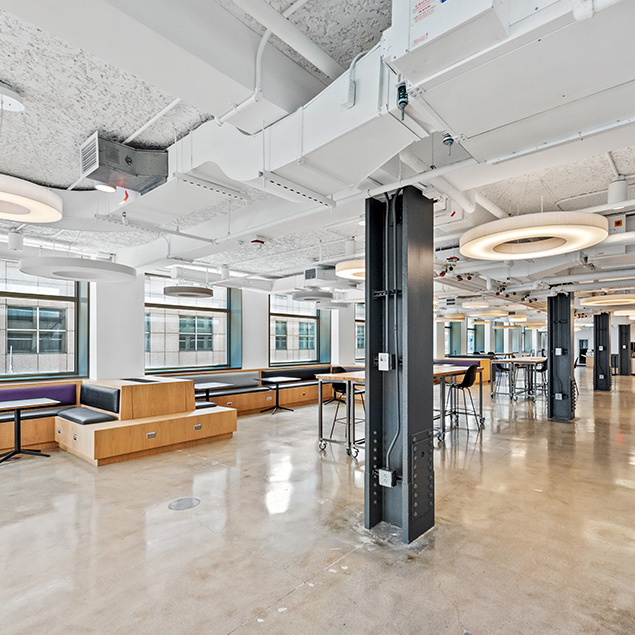 Modern open office space with polished floors, exposed ceilings, large windows, built-in wooden seating, high tables with stools, and circular ceiling lights, creating a bright and airy environment.
