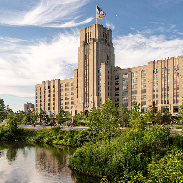 A large, historic stone building with a central tower and an American flag on top stands behind lush greenery and a pond under a blue sky with wispy clouds.