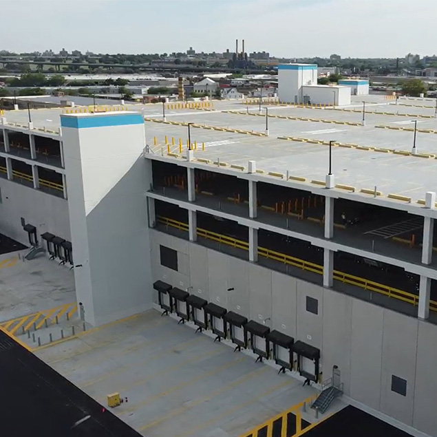 A large, multi-level warehouse building with loading docks on the ground floor and parking areas above, surrounded by a cityscape and industrial structures in the background.