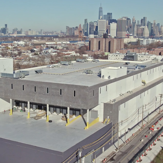 A large, modern industrial warehouse with loading docks, surrounded by a cityscape and high-rise buildings in the background under a clear sky.