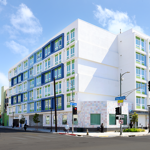 A modern, multi-story apartment building with blue, green, and white accents sits on a sunny street corner. People walk on the sidewalk, and street signs mark the intersection of 6th Street and San Julian Street.