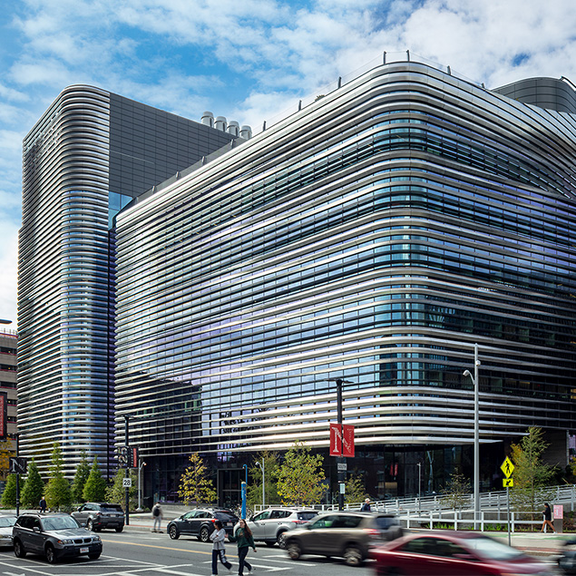 A modern building with curved, horizontal metal bands and large glass windows stands at a busy street corner, with cars and pedestrians passing by and a few trees lining the sidewalk.