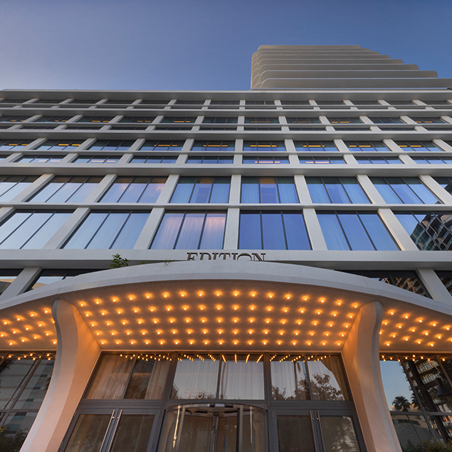 Low-angle view of a modern hotel building with large windows and a curved entrance canopy lit by many small lights. The word EDITION is partially visible above the doorway.