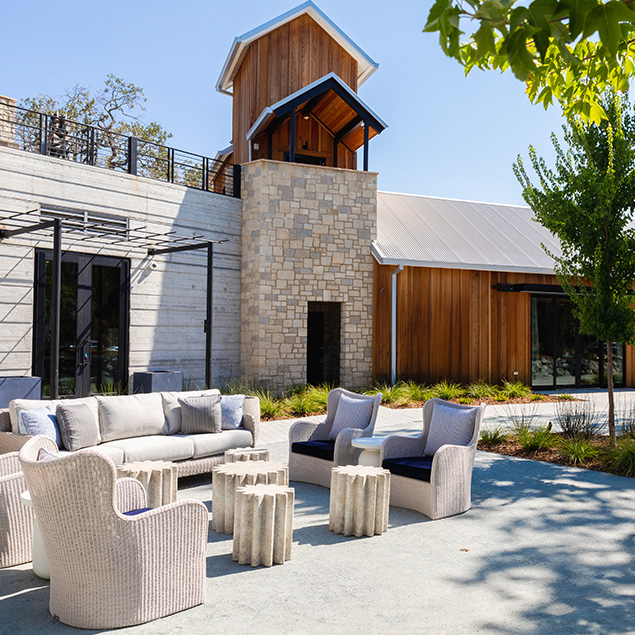 Outdoor patio with cushioned chairs, sofas, and small tables arranged in front of a modern building with wood and stone exterior. Trees and landscaped plants surround the seating area on a sunny day.