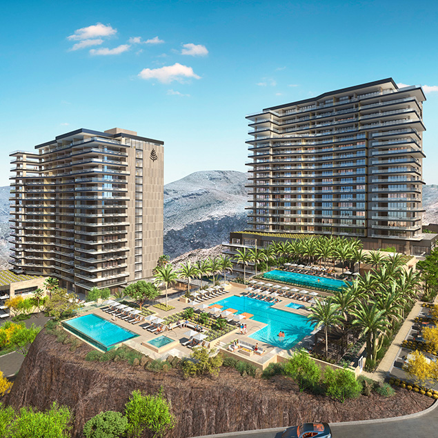 A modern resort complex features two tall buildings and a central outdoor area with swimming pools, lounge chairs, and palm trees, set against a backdrop of rocky hills under a clear blue sky.