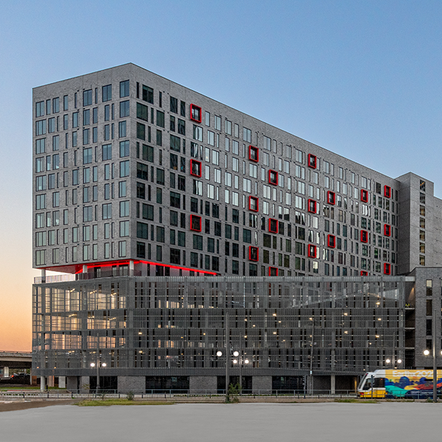 A modern multi-story building with a grid-like facade, red accents around some windows, and a glass lower section. A colorful tram passes in front of the building at dusk.