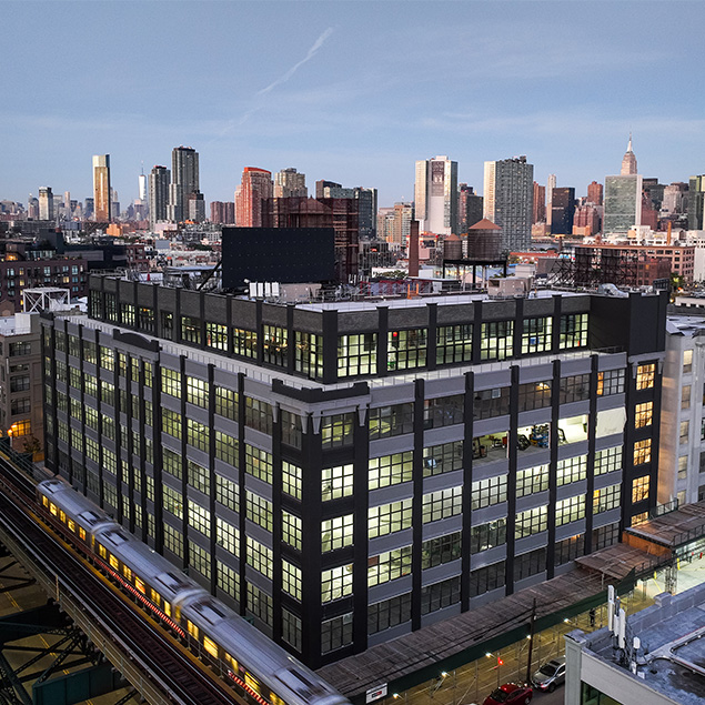 A large, modern office building with many windows is shown at dusk in an urban area. A subway train is passing by on elevated tracks in the foreground, with a city skyline in the background.