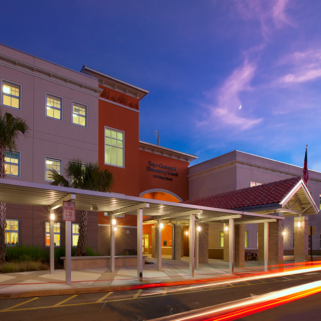 A modern school building glows with lights at dusk. The entrance features a covered walkway, palm trees, and a sign reading Hope Centennial Elementary School. Light trails from passing cars are visible in the foreground.