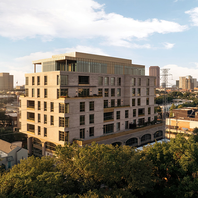 A modern, multi-story building with large windows and balconies stands amidst trees, with a city skyline and partly cloudy sky in the background.