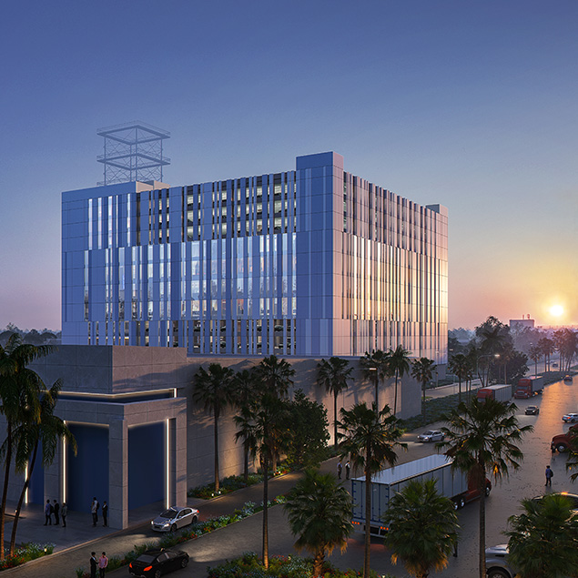 A modern, multi-story building with reflective glass windows stands along a palm-lined street at sunset. Cars and trucks are parked nearby, and small groups of people are gathered by the building’s entrance.