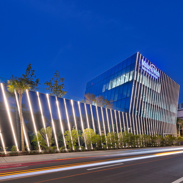 A modern glass building with the Make-A-Wish logo at dusk, illuminated by vertical exterior lights, with palm trees and light trails from passing vehicles in the foreground.