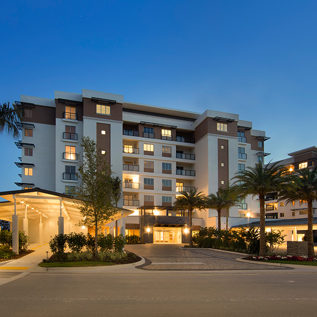 A modern multi-story hotel building is illuminated at dusk, with glowing windows, palm trees, and landscaped gardens in front of the entrance under a covered driveway.