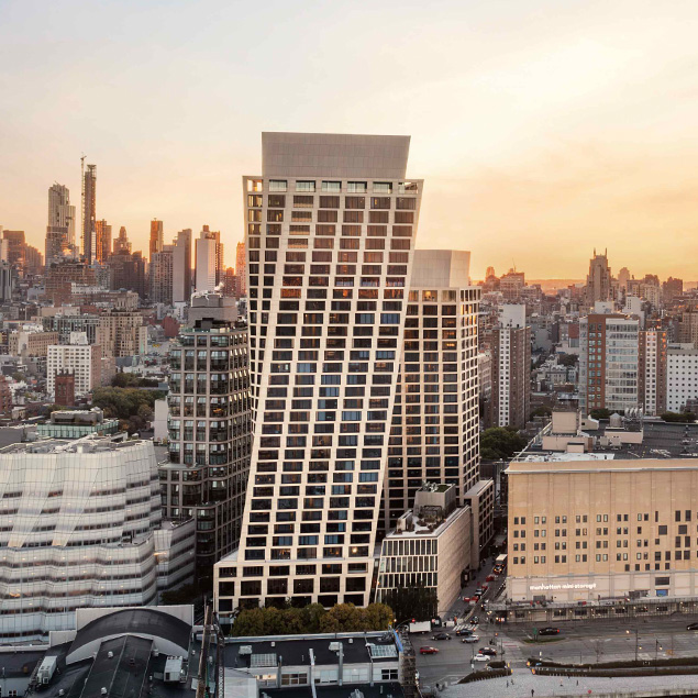 A modern skyscraper with a distinctive slanted design stands among other buildings in a cityscape at sunset, with warm light illuminating the scene and the skyline visible in the background.
