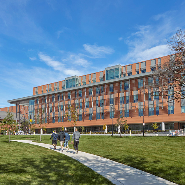 A group of four people walk along a curved path on a grassy lawn toward a modern, multi-story brick and glass building under a clear blue sky.