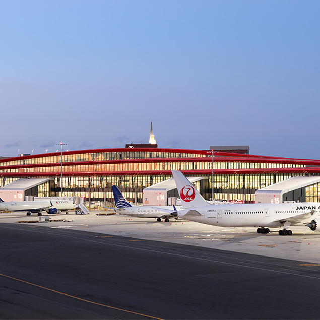 Several airplanes are parked at airport gates in front of a modern terminal building with large glass windows and red accents at dusk.